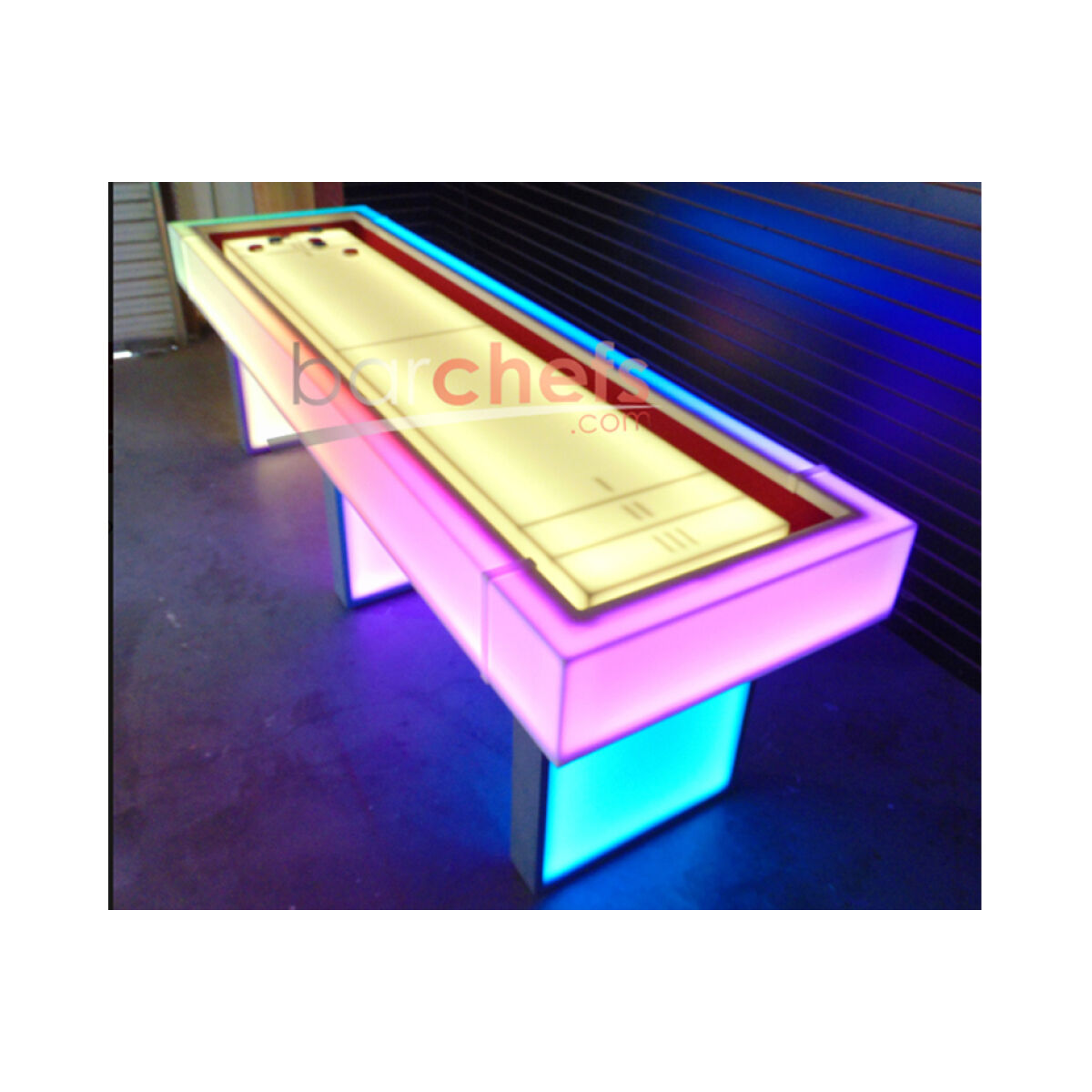 An illuminated shuffleboard table featuring a wide, smooth playing surface, bordered with vibrant pink, blue, and purple lighting. The surface includes a red scoring area and multiple holes for gameplay. The backdrop is dark, emphasizing the table's colorful illumination.